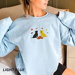 Woman in a Light Blue crewneck sweatshirt embroidered with a black Labrador and a yellow Labrador sitting by three pumpkins under falling autumn leaves