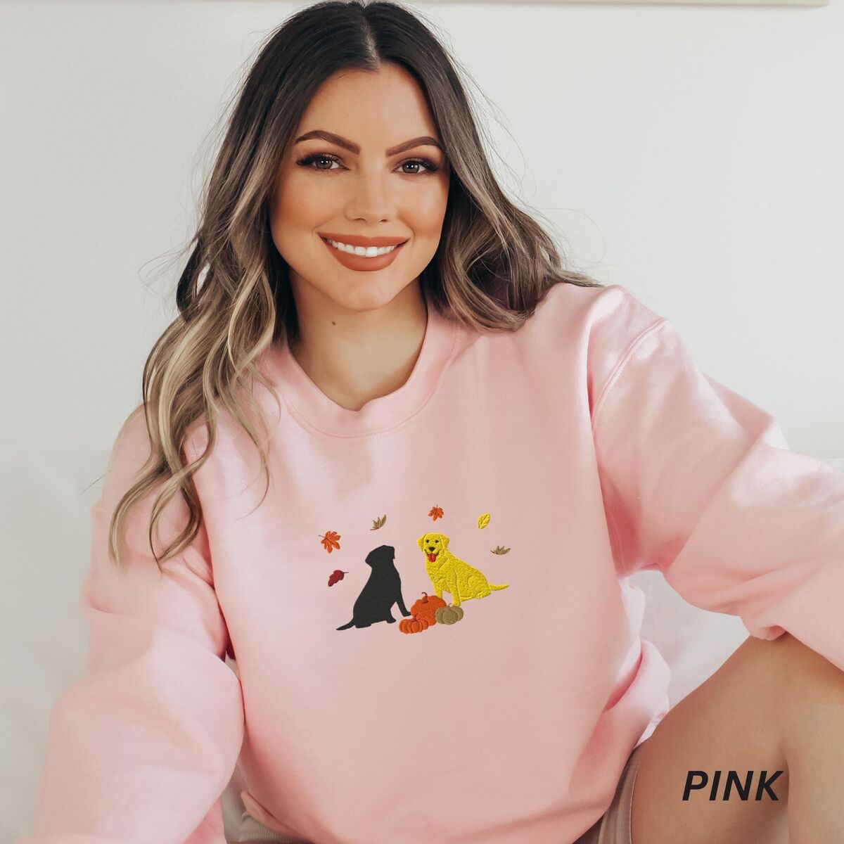 Black and Yellow Lab Fall Sweatshirt - Light Pink Smiling woman in a Light Pink crewneck sweatshirt embroidered with a black Labrador and a yellow Labrador sitting by three pumpkins under falling autumn leaves