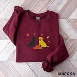Maroon crewneck sweatshirt laid flat on herringbone wood, embroidered with a black Labrador and a yellow Labrador sitting by three pumpkins under falling autumn leaves