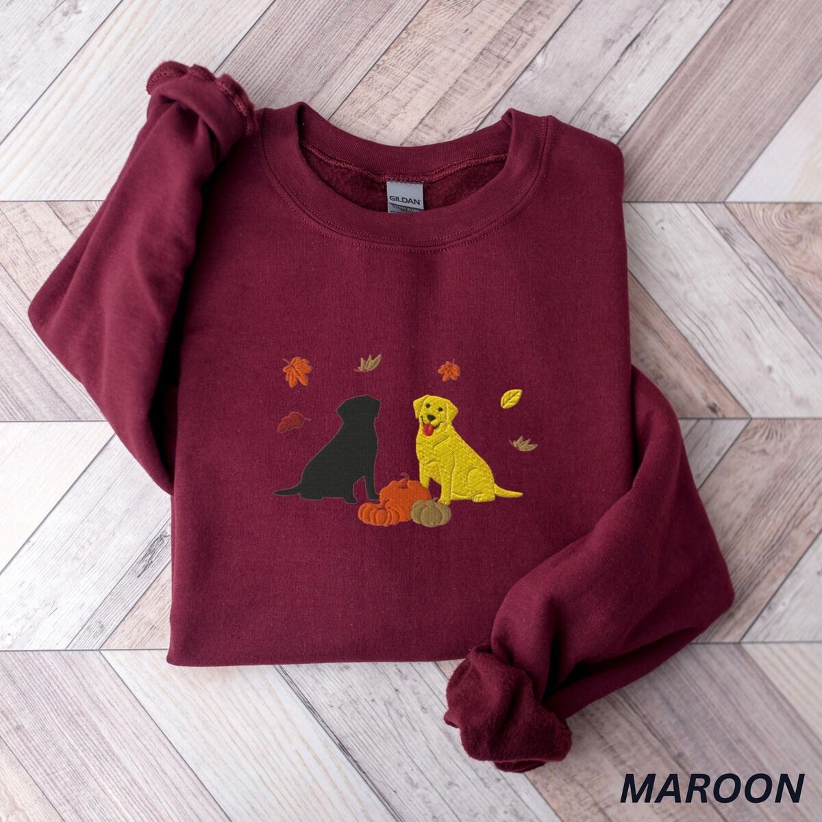 Black and Yellow Lab Fall Sweatshirt – Maroon Maroon crewneck sweatshirt laid flat on herringbone wood, embroidered with a black Labrador and a yellow Labrador sitting by three pumpkins under falling autumn leaves