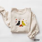 Sand crewneck sweatshirt laid flat on white, embroidered with a black Labrador and a yellow Labrador sitting by three pumpkins under falling autumn leaves