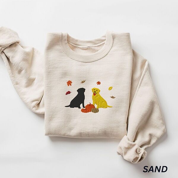 Sand crewneck sweatshirt laid flat on white, embroidered with a black Labrador and a yellow Labrador sitting by three pumpkins under falling autumn leaves