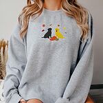 Woman in a Sport Grey crewneck sweatshirt embroidered with a black Labrador and a yellow Labrador sitting by three pumpkins under falling autumn leaves