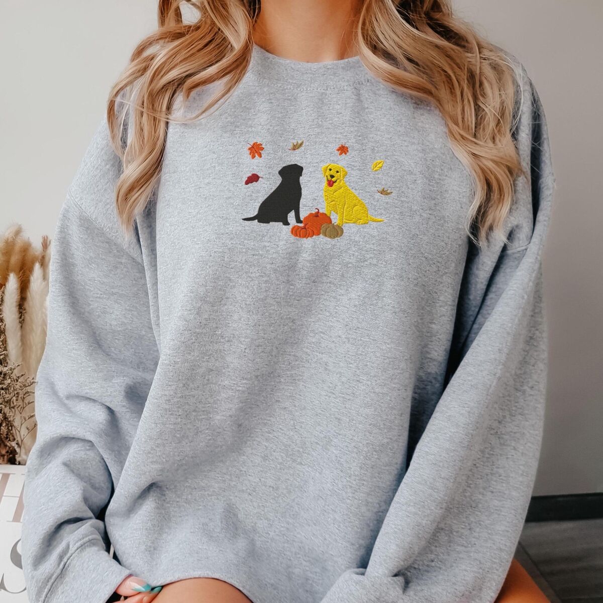 Black and Yellow Lab Fall Sweatshirt - Sport Grey Woman in a Sport Grey crewneck sweatshirt embroidered with a black Labrador and a yellow Labrador sitting by three pumpkins under falling autumn leaves