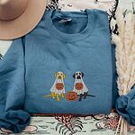 Indigo Blue crewneck sweatshirt laid flat with a brown hat and pampas grass, embroidered with a yellow lab and a black lab sitting in white ghost sheets, each hugging a carved jack-o'-lantern