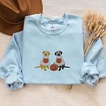 Light Blue crewneck sweatshirt laid flat with a brown hat and wheat stems, embroidered with a yellow lab and a black lab sitting in white ghost sheets, each hugging a carved jack-o'-lantern