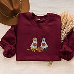 Maroon crewneck sweatshirt laid flat with a brown hat and wheat stems, embroidered with a yellow lab and a black lab sitting in white ghost sheets, each hugging a carved jack-o'-lantern