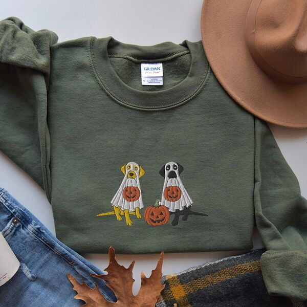Military Green crewneck sweatshirt laid flat beside jeans, a plaid scarf and autumn leaves, embroidered with a yellow lab and a black lab sitting in white ghost sheets, each hugging a carved jack-o'-lantern