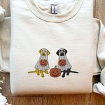Sand crewneck sweatshirt laid flat on a wood floor, embroidered with a yellow lab and a black lab sitting in white ghost sheets, each hugging a carved jack-o'-lantern