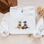 White crewneck sweatshirt laid flat with a brown hat and wheat stems, embroidered with a yellow lab and a black lab sitting in white ghost sheets, each hugging a carved jack-o'-lantern