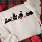 Close-up of four embroidered black cats with a Santa hat, string lights, a present and an ornament on a cream sweatshirt