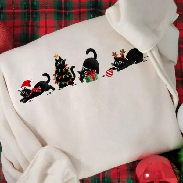 Close-up of four embroidered black cats with a Santa hat, string lights, a present and an ornament on a cream sweatshirt
