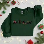Forest green Christmas sweatshirt with a row of embroidered black holiday cats, styled with gifts and lights