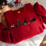 Red sweatshirt with embroidered black cats in a Santa hat, Christmas lights, a gift box and antlers