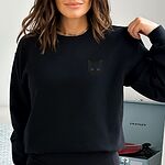 Woman wearing a Black crewneck sweatshirt beside a record player, embroidered with a solid black cat face with bright eyes on the left chest