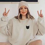 Smiling woman in a beanie making peace signs in a sand crewneck sweatshirt, embroidered with a solid black cat face with bright eyes on the left chest