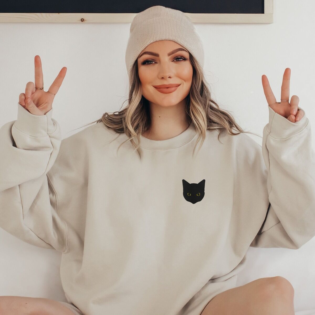 Black Cat Face Sweatshirt - worn in sand Smiling woman in a beanie making peace signs in a sand crewneck sweatshirt, embroidered with a solid black cat face with bright eyes on the left chest