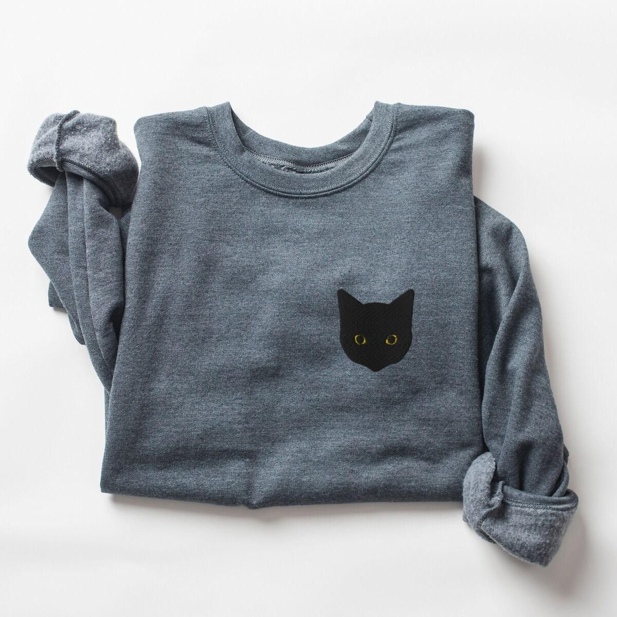 Black Cat Face Sweatshirt – Dark Heather Dark Heather crewneck sweatshirt laid flat with rolled sleeves, embroidered with a solid black cat face with yellow eyes on the left chest