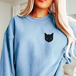 Woman wearing a Light Blue crewneck sweatshirt, embroidered with a solid black cat face with blue eyes on the left chest