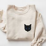 Sand crewneck sweatshirt laid flat with a rolled sleeve, embroidered with a solid black cat face with blue eyes on the left chest