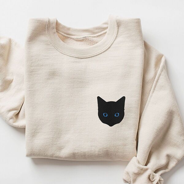 Sand crewneck sweatshirt laid flat with a rolled sleeve, embroidered with a solid black cat face with blue eyes on the left chest