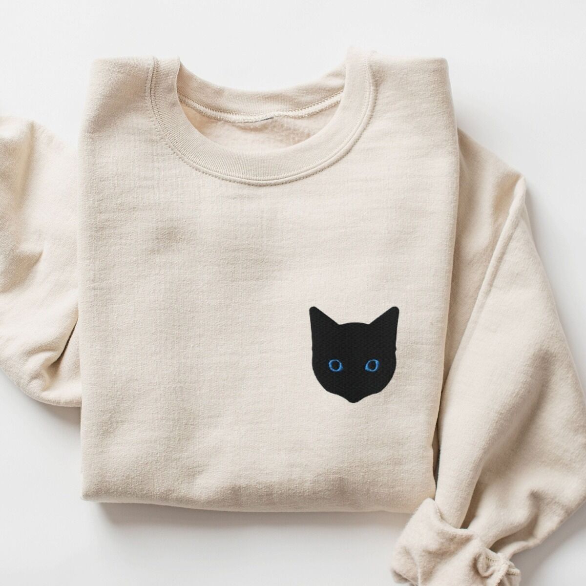 Black Cat Face Sweatshirt – Sand Sand crewneck sweatshirt laid flat with a rolled sleeve, embroidered with a solid black cat face with blue eyes on the left chest