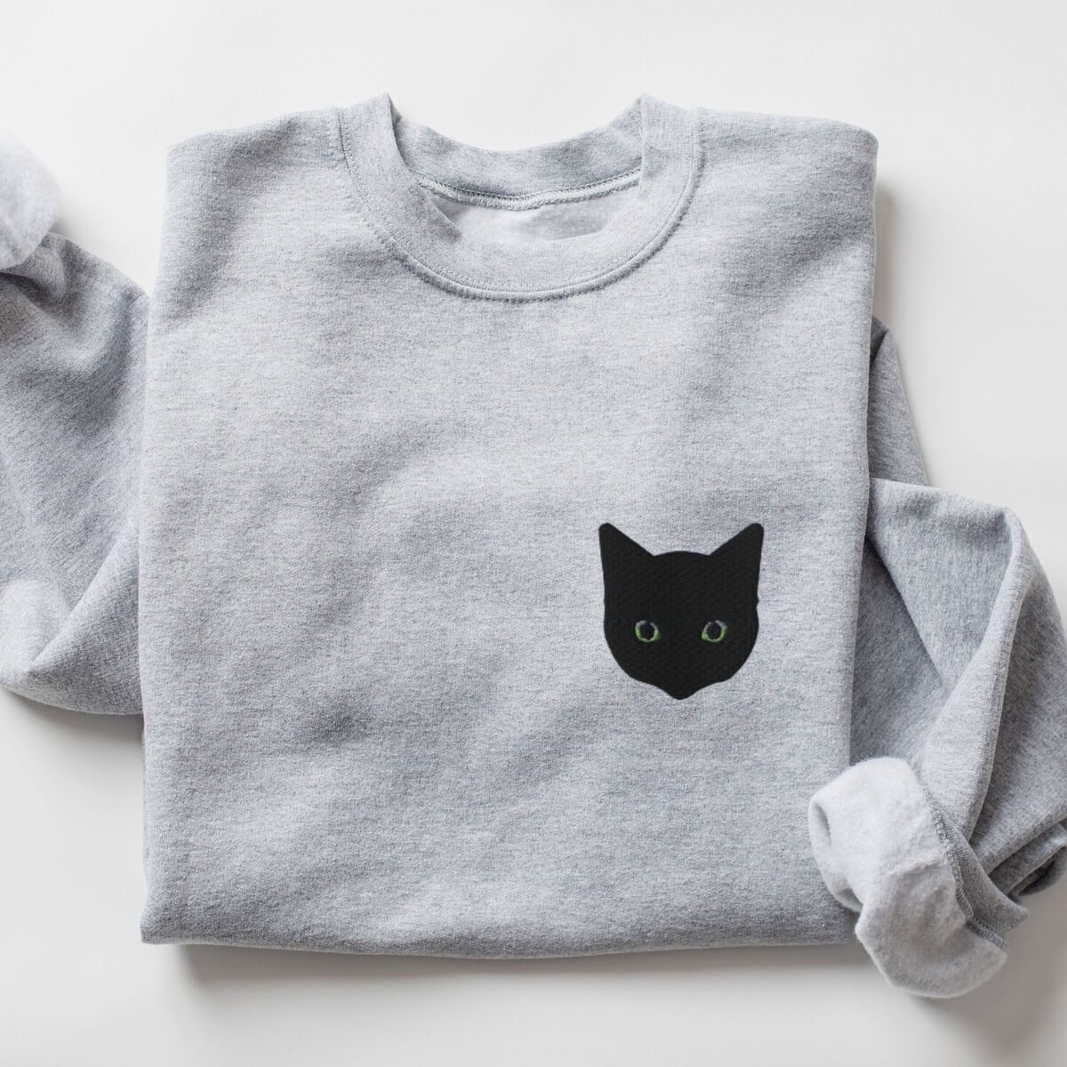Black Cat Face Sweatshirt - Sport Grey Sport Grey crewneck sweatshirt laid flat with a rolled sleeve, embroidered with a solid black cat face with green eyes on the left chest
