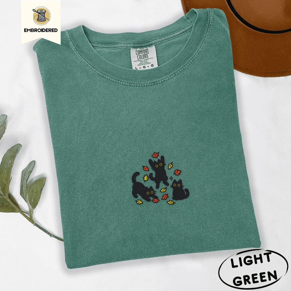 Black Cat Fall Shirt – Light Green Light Green Comfort Colors T-shirt embroidered with three black cats pouncing, leaping and sitting among falling red, orange and yellow autumn leaves