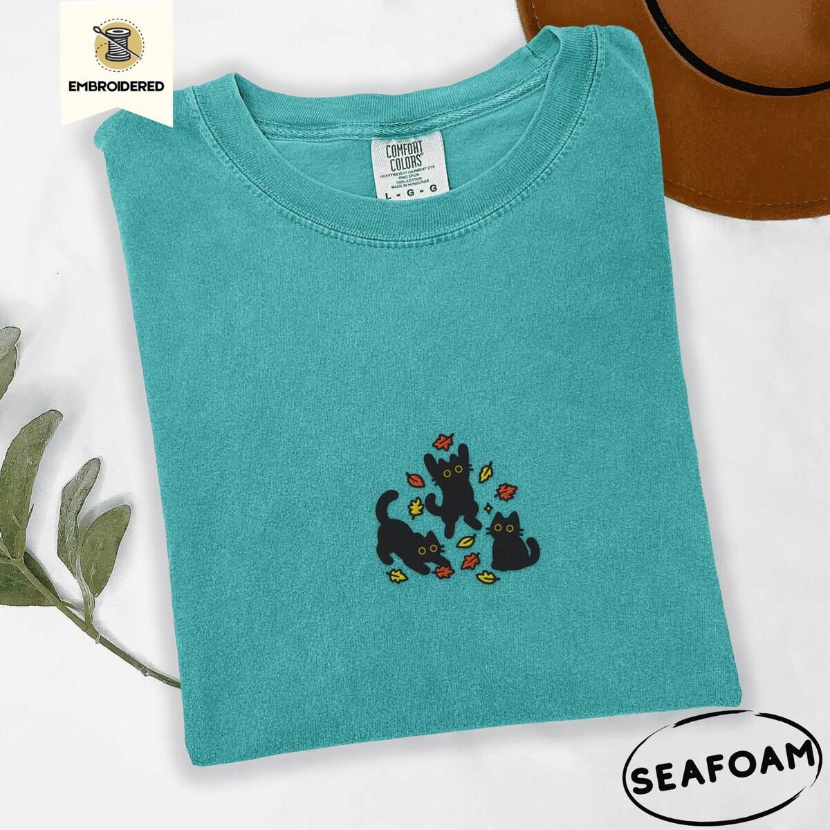 Black Cat Fall Shirt – Seafoam Seafoam Comfort Colors T-shirt embroidered with three black cats pouncing, leaping and sitting among falling red, orange and yellow autumn leaves