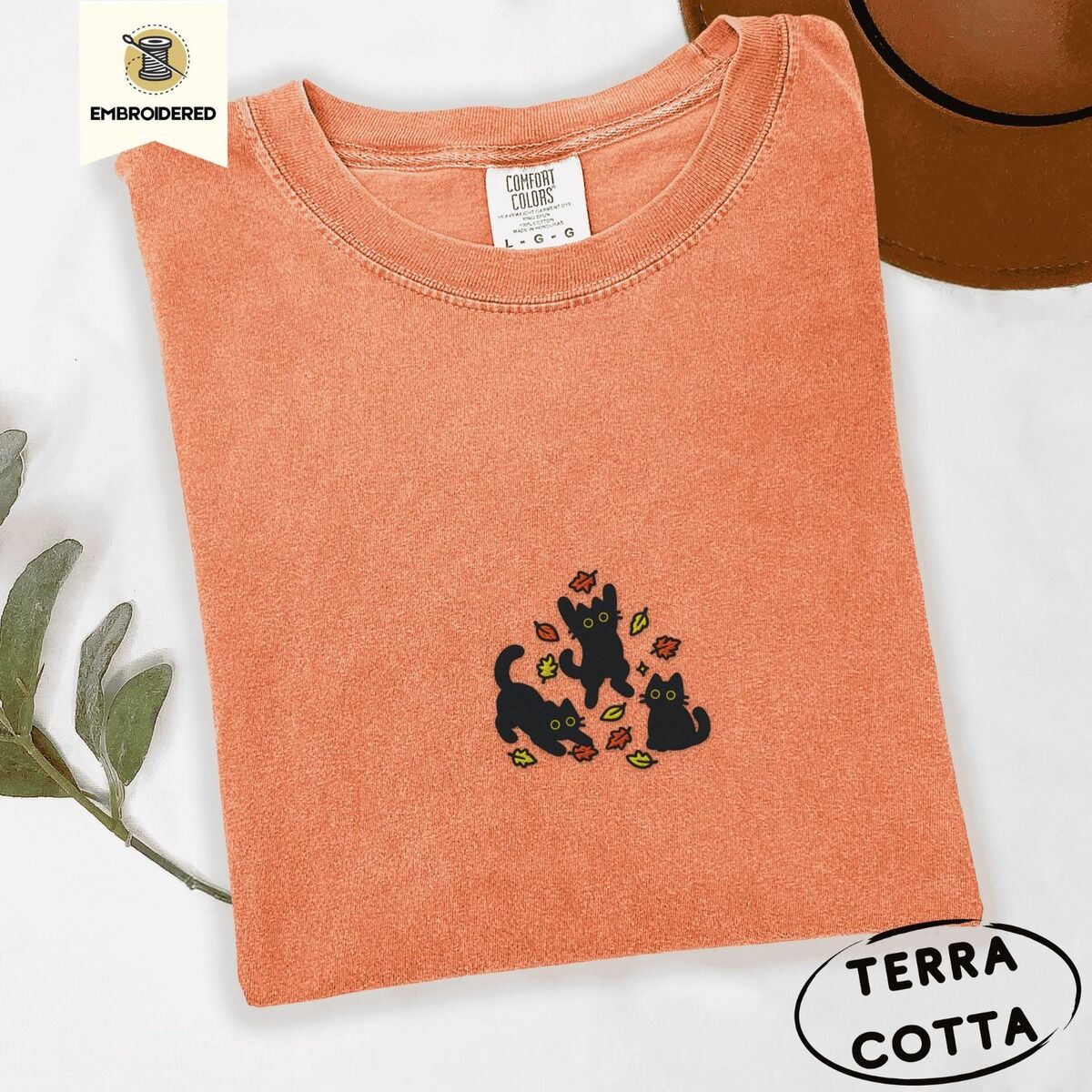 Black Cat Fall Shirt – Terracotta Terracotta Comfort Colors T-shirt embroidered with three black cats pouncing, leaping and sitting among falling red, orange and yellow autumn leaves