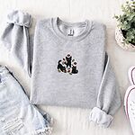 Ash crewneck sweatshirt folded with denim shorts, white sneakers and a potted plant, embroidered with three black cats pouncing, leaping and sitting among falling red, orange and yellow autumn leaves