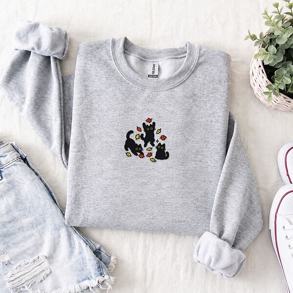 Ash crewneck sweatshirt folded with denim shorts, white sneakers and a potted plant, embroidered with three black cats pouncing, leaping and sitting among falling red, orange and yellow autumn leaves