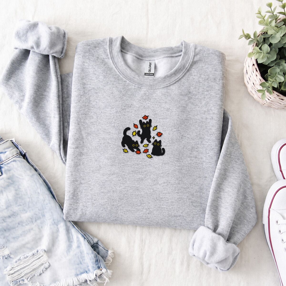 Black Cat Fall Sweatshirt – Ash Ash crewneck sweatshirt folded with denim shorts, white sneakers and a potted plant, embroidered with three black cats pouncing, leaping and sitting among falling red, orange and yellow autumn leaves