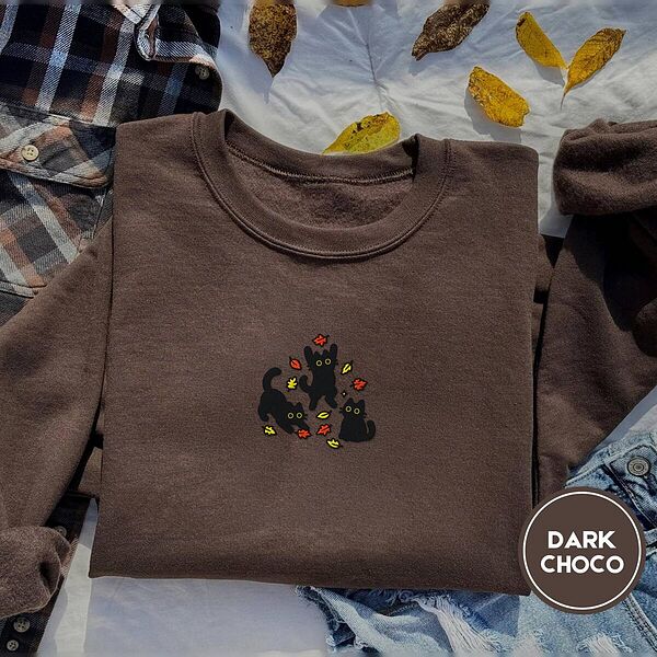 Dark Chocolate crewneck sweatshirt on a plaid flannel shirt with yellow autumn leaves and denim shorts, embroidered with three black cats pouncing, leaping and sitting among falling red, orange and yellow autumn leaves