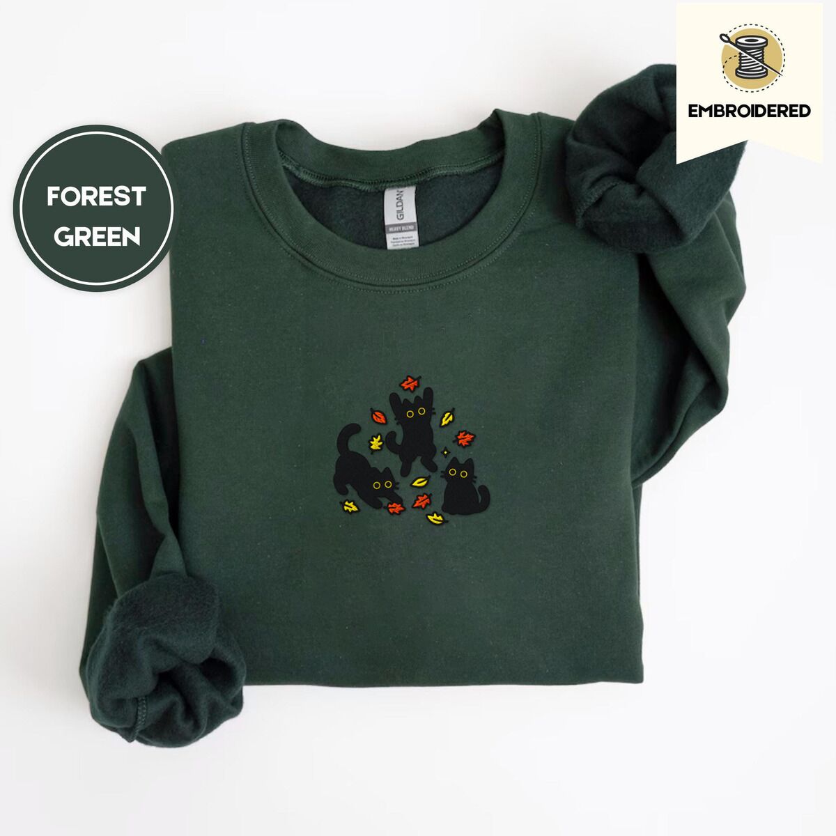 Black Cat Fall Sweatshirt – Forest Green Forest Green crewneck sweatshirt folded on a white background, embroidered with three black cats pouncing, leaping and sitting among falling red, orange and yellow autumn leaves