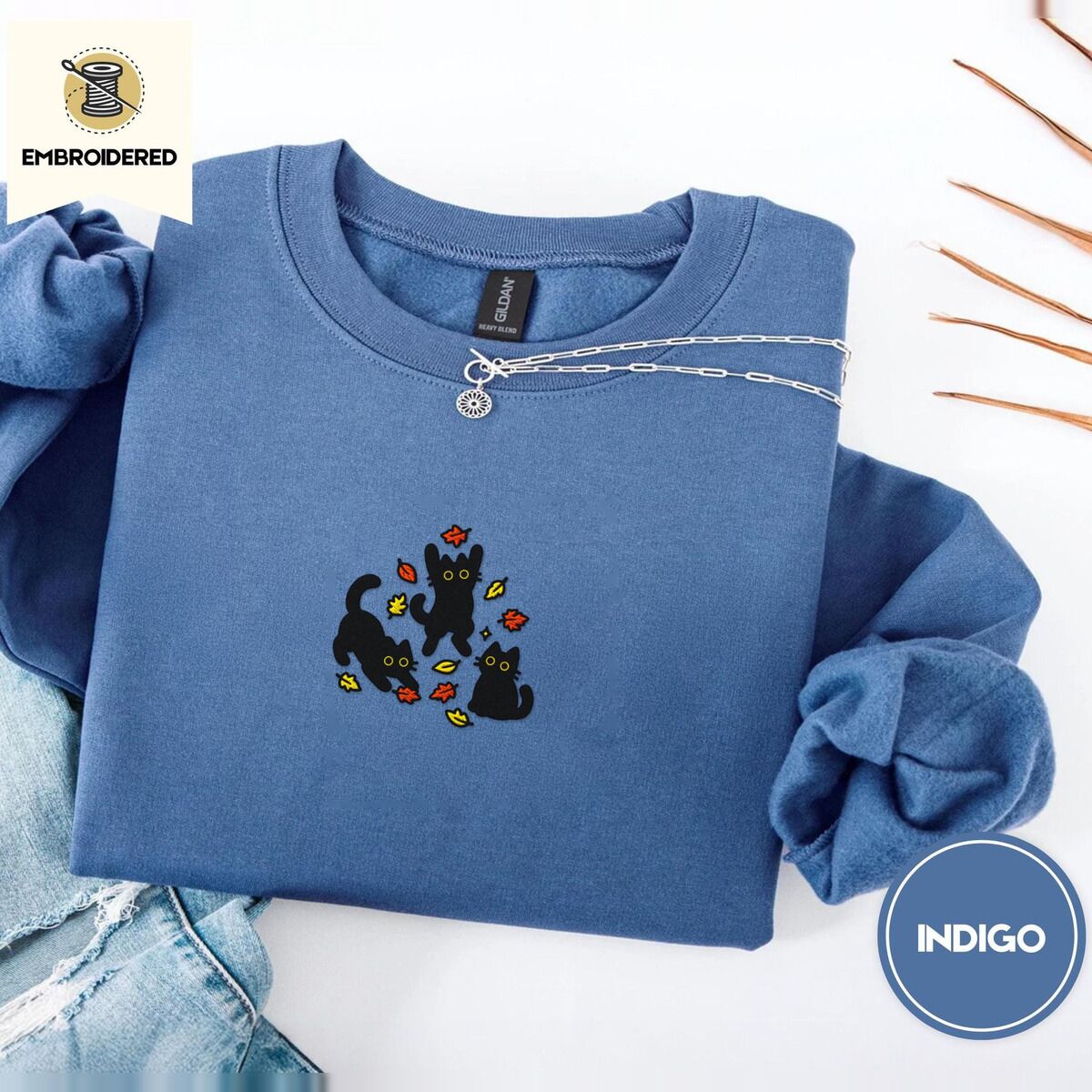 Black Cat Fall Sweatshirt – Indigo Blue Indigo Blue crewneck sweatshirt folded with light jeans and dried twigs, embroidered with three black cats pouncing, leaping and sitting among falling red, orange and yellow autumn leaves