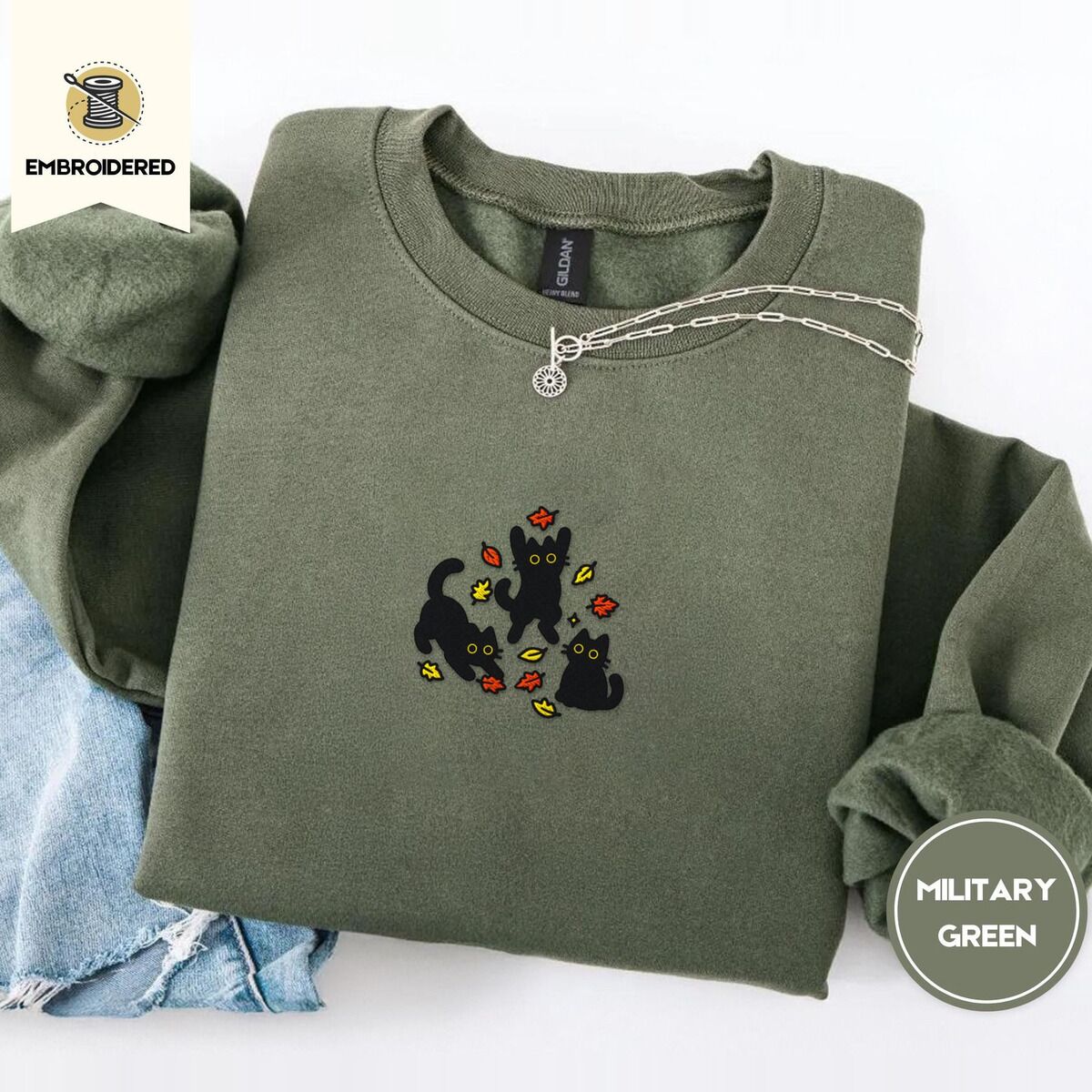 Black Cat Fall Sweatshirt – Military Green Military Green crewneck sweatshirt folded with light jeans and a silver chain necklace, embroidered with three black cats pouncing, leaping and sitting among falling red, orange and yellow autumn leaves