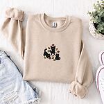 Sand crewneck sweatshirt folded with denim shorts, white sneakers and a potted plant, embroidered with three black cats pouncing, leaping and sitting among falling red, orange and yellow autumn leaves