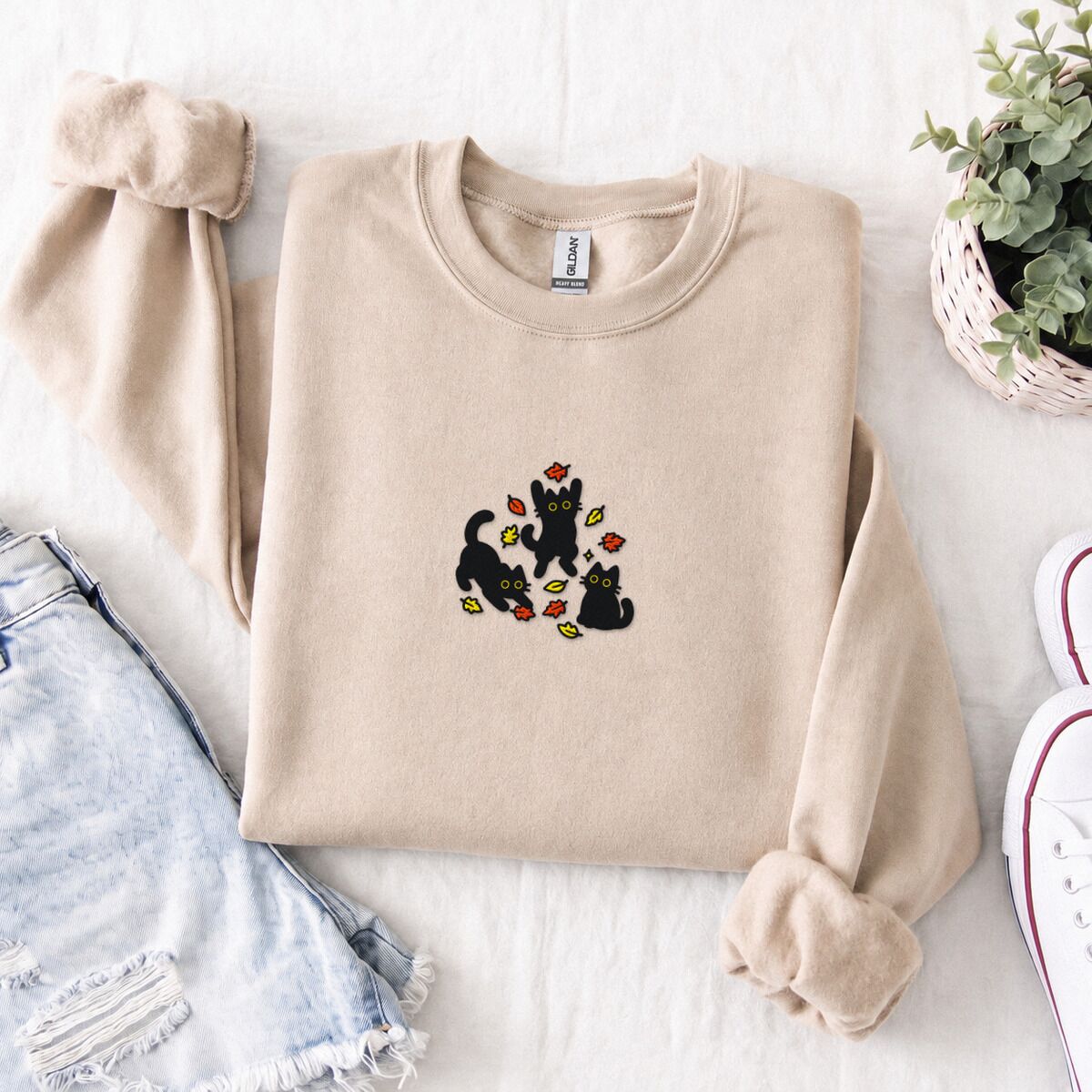 Black Cat Fall Sweatshirt – Sand Sand crewneck sweatshirt folded with denim shorts, white sneakers and a potted plant, embroidered with three black cats pouncing, leaping and sitting among falling red, orange and yellow autumn leaves