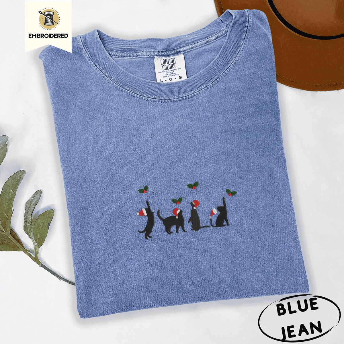 Black Cat Santa Hat Shirt – Blue Jean Blue Jean Comfort Colors T-shirt embroidered with four black cat silhouettes in Santa hats reaching for holly across the chest