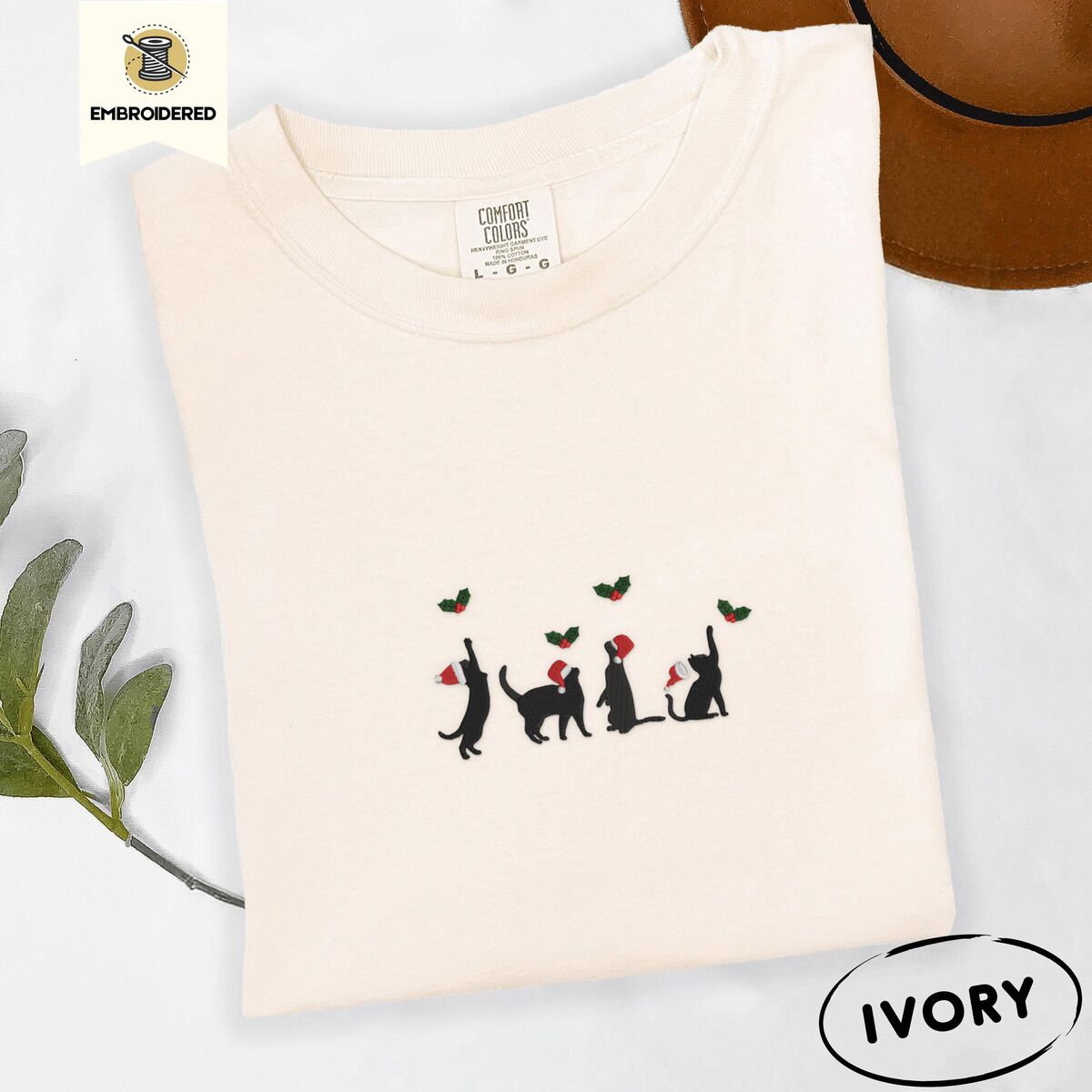Black Cat Santa Hat Shirt – Ivory Ivory Comfort Colors T-shirt embroidered with four black cat silhouettes in Santa hats reaching for holly across the chest