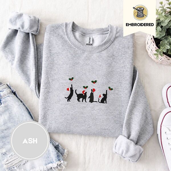 Ash crewneck sweatshirt folded with denim shorts, white sneakers and a potted plant, embroidered with four black cat silhouettes in Santa hats reaching for holly across the chest