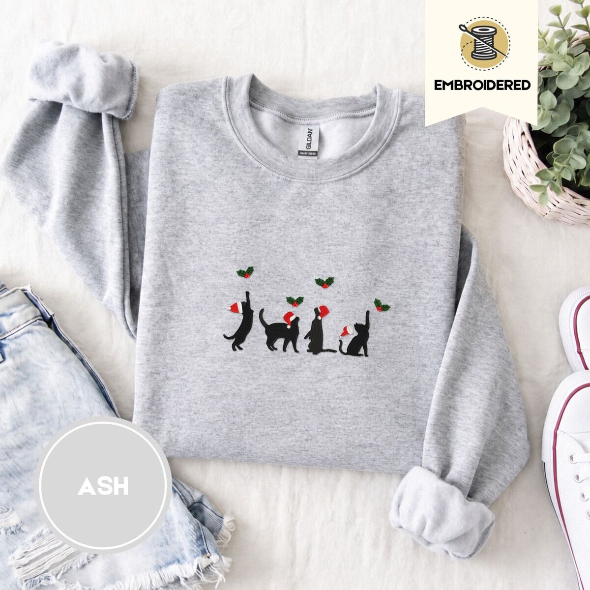 Black Cat Santa Hat Sweatshirt – Ash Ash crewneck sweatshirt folded with denim shorts, white sneakers and a potted plant, embroidered with four black cat silhouettes in Santa hats reaching for holly across the chest