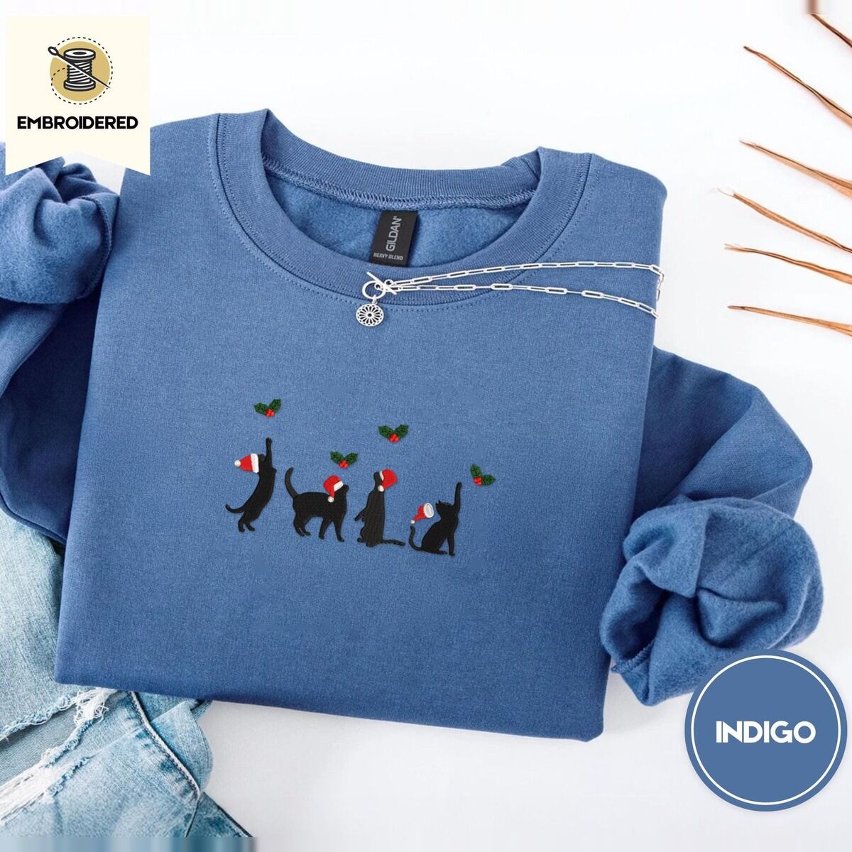 Black Cat Santa Hat Sweatshirt – Indigo Blue Indigo Blue crewneck sweatshirt folded with light jeans and dried twigs, embroidered with four black cat silhouettes in Santa hats reaching for holly across the chest