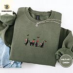 Military Green crewneck sweatshirt folded with light jeans and a silver chain necklace, embroidered with four black cat silhouettes in Santa hats reaching for holly across the chest