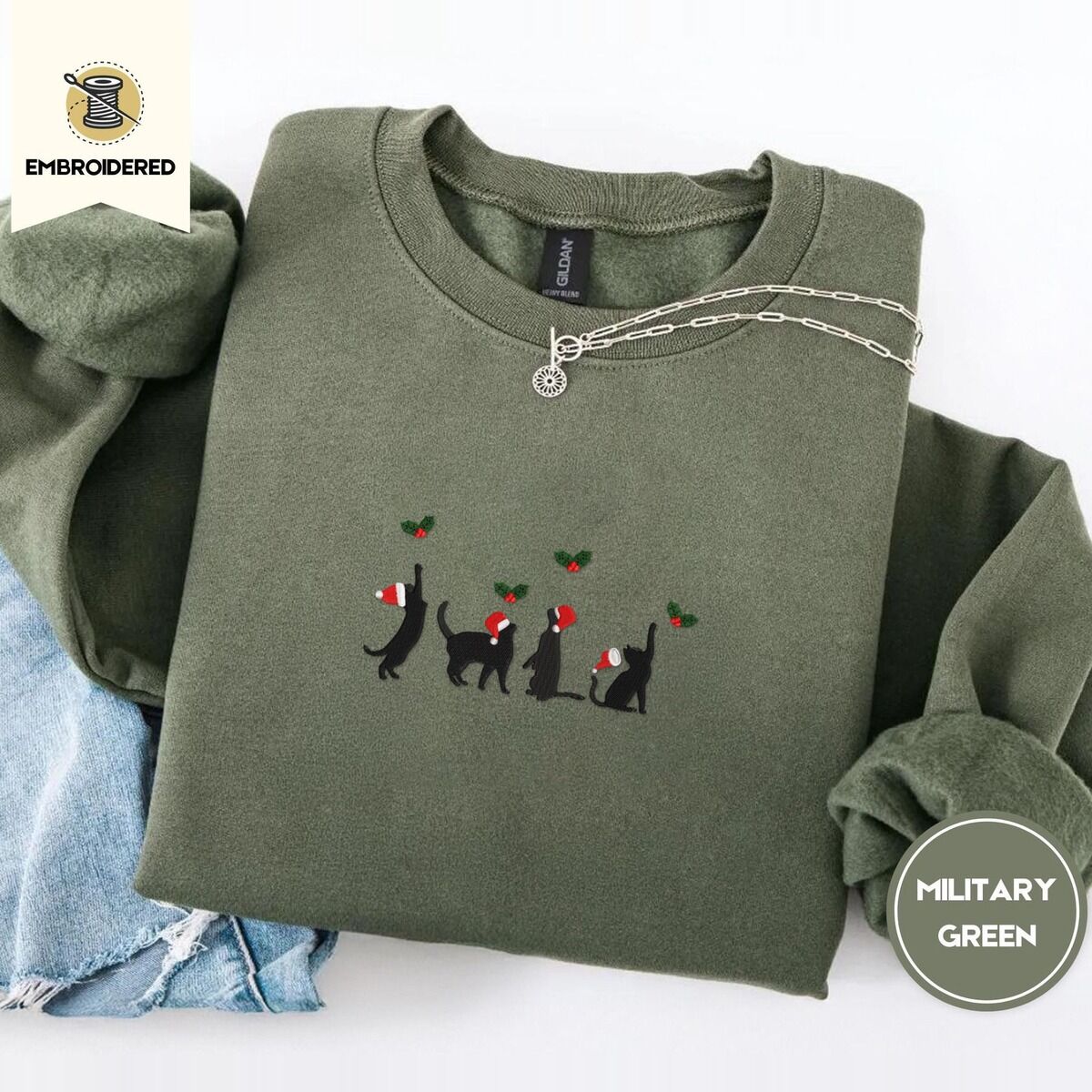 Black Cat Santa Hat Sweatshirt – Military Green Military Green crewneck sweatshirt folded with light jeans and a silver chain necklace, embroidered with four black cat silhouettes in Santa hats reaching for holly across the chest