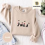 Sand crewneck sweatshirt folded with denim shorts, white sneakers and a potted plant, embroidered with four black cat silhouettes in Santa hats reaching for holly across the chest