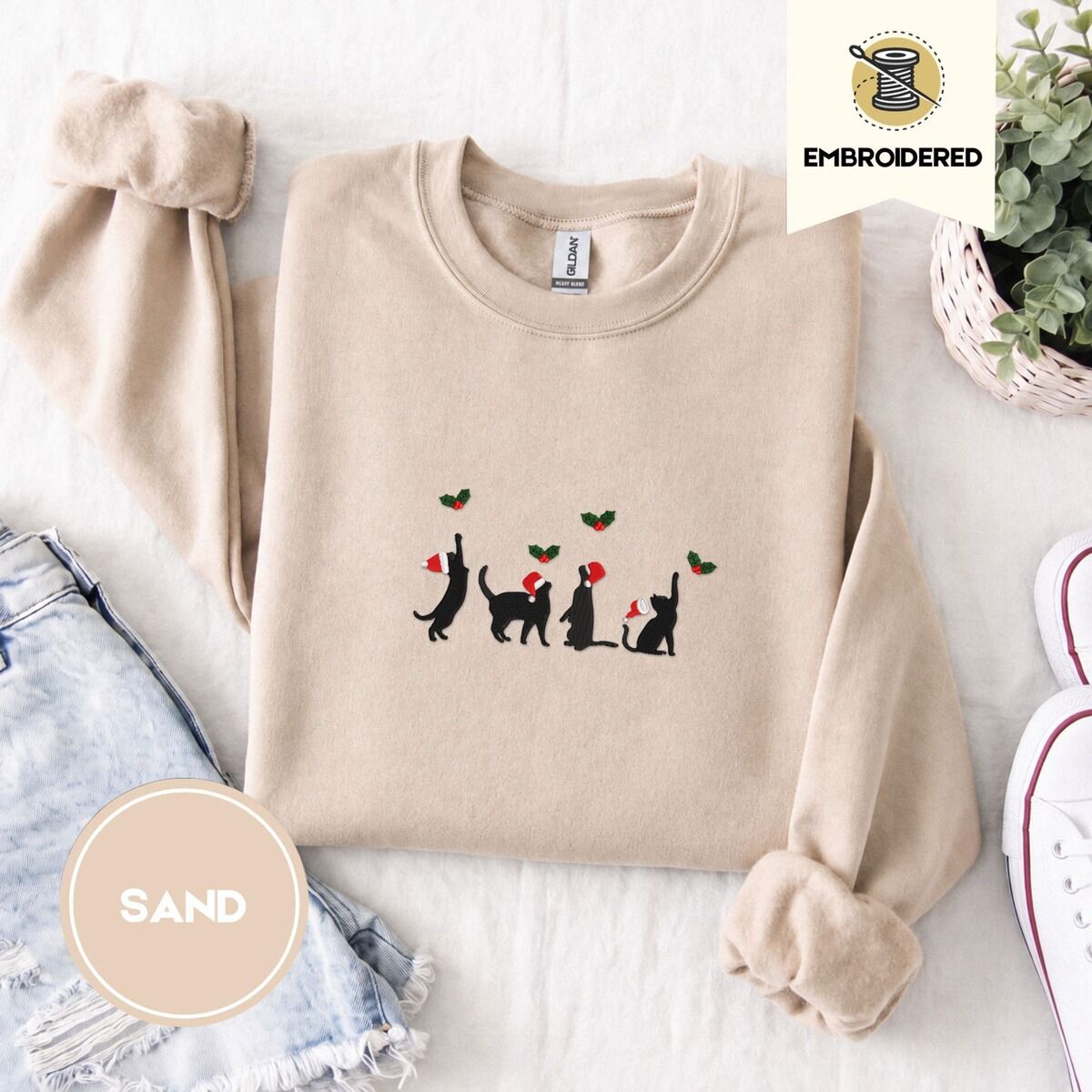 Black Cat Santa Hat Sweatshirt – Sand Sand crewneck sweatshirt folded with denim shorts, white sneakers and a potted plant, embroidered with four black cat silhouettes in Santa hats reaching for holly across the chest