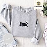 Ash crewneck sweatshirt folded with denim shorts, white sneakers and a potted plant, embroidered with a black cat lounging with a glass of pink wine in the center of the chest