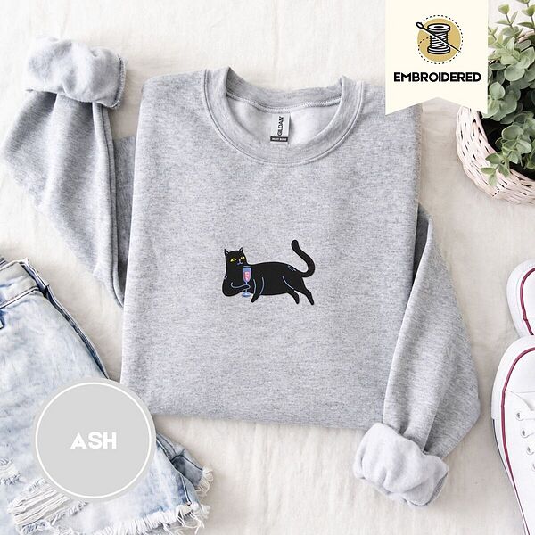 Ash crewneck sweatshirt folded with denim shorts, white sneakers and a potted plant, embroidered with a black cat lounging with a glass of pink wine in the center of the chest
